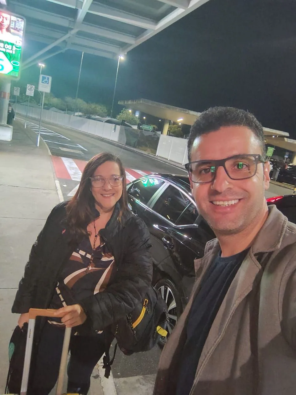 Cliente com a mala ao lado do motorista da TEVO no Aeroporto de Confins, à noite, junto ao carro executivo