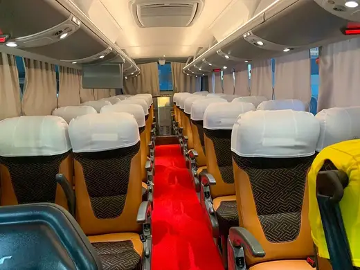 Interior de ônibus com poltronas caramelo, capas brancas e corredor com tapete vermelho