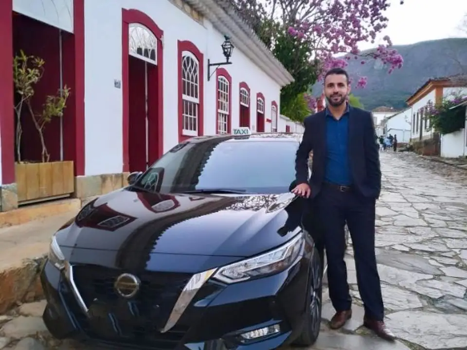 Motorista de terno ao lado do táxi executivo preto em rua de pedra de Tiradentes, com ipê florido